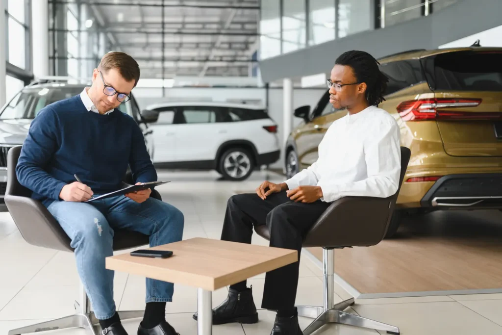 Two men discussing a car invoice at a dealership surrounded by new vehicles.
