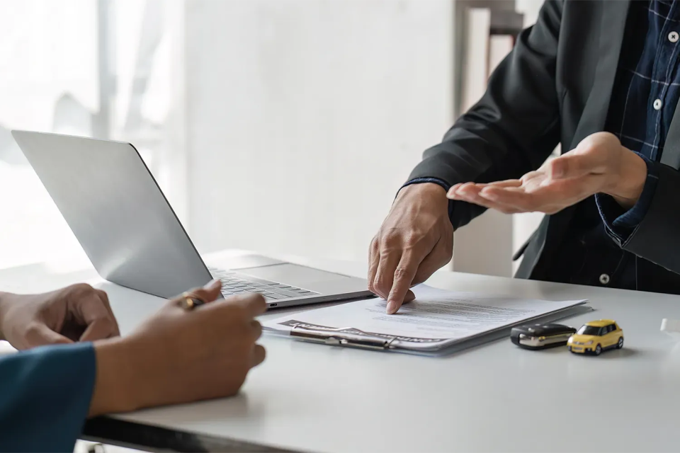 Customer and car dealer discussing invoice price details with contract and toy cars on a desk