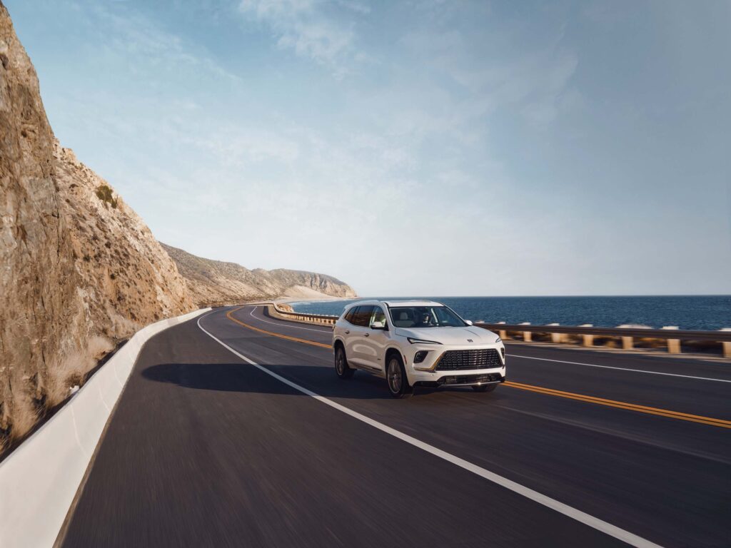 White Buick Enclave driving along a coastal highway beside the ocean, featured in our Buick Enclave Review.