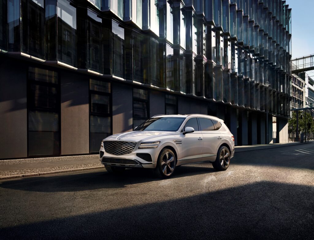 White Genesis GV80 luxury SUV parked on a city street beside modern glass buildings for a Genesis GV80 Review.