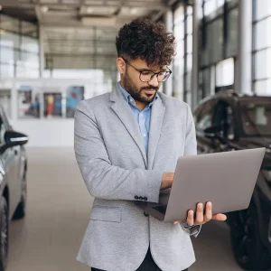 Man using a laptop at a car dealership to perform a dealer invoice price lookup on new vehicles