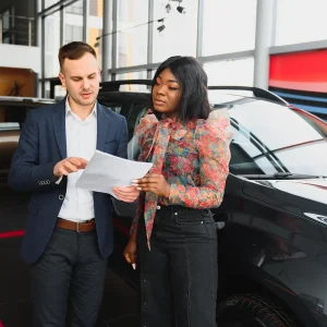 Dealer and customer discussing invoice pricing for cars while reviewing documents beside a new vehicle.